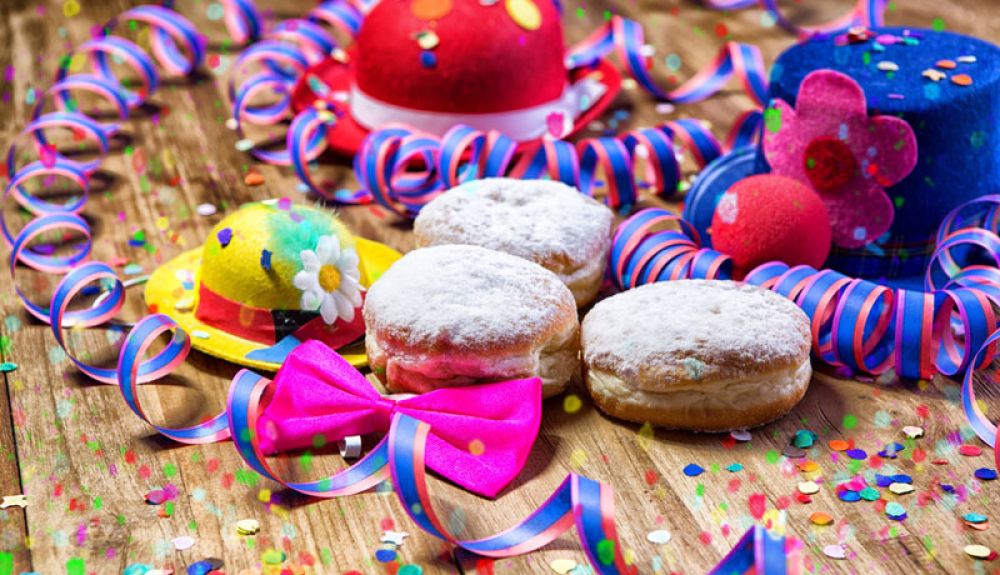 Das Foto zeigt drei gefüllte Krapfen auf einem Holztisch mit bunten Hüten, Konfetti und Luftschlangen.